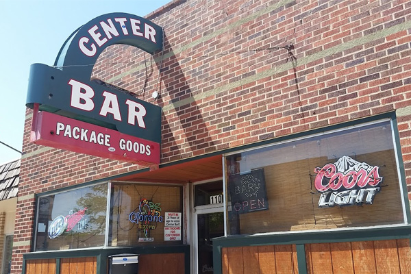 A black sign with “Center Bar” in white text hangs above a red sign for “Package” and “Goods,” with a light for “Coors Light” in the window of a brick wall.
