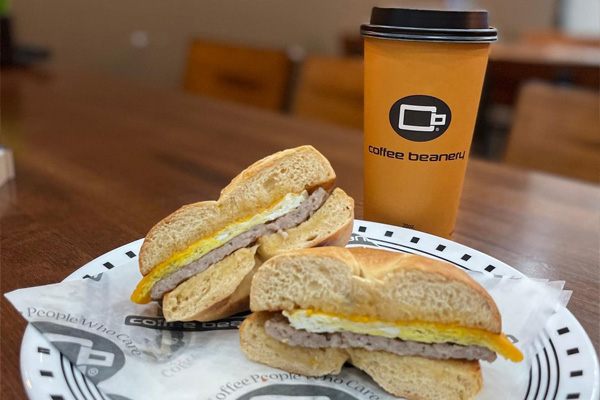 A Coffee Beanery to-go cup of coffee with a breakfast sandwich served on Coffee Beanery’s paper, ready to eat on a black and white plate.