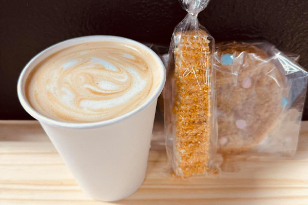 Tenille’s coffee shop, Teak Coffee, serves a to-go latte paired with two packaged pastries on a wooden table.