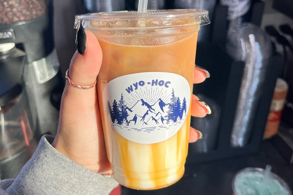 A girl with a manicure holds a plastic to-go cup of creamy coffee from Wyoming House of Coffee, a specialty Wyoming coffee shop. The order is served with a white and blue mountains sticker with trees, “WYO-HOC” printed at the top.