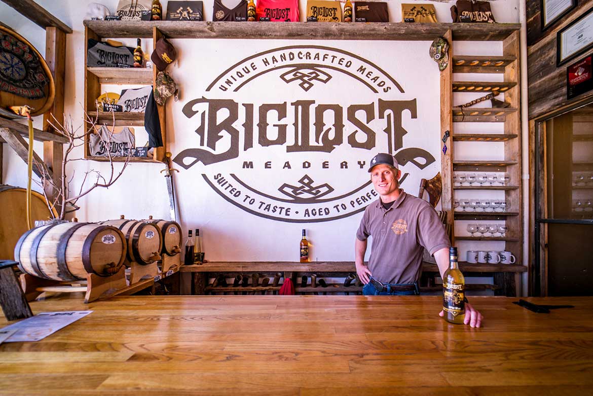 Sam Clikeman, owner of Big Lost Meadery, holds a bottle of handcrafted mead in front of a big sign that advertises, “Big Lost Meadery: Crafted to Taste. Aged to perfection.”